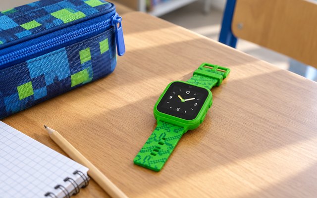 Kids' smartwatch, lying on a school desk beside a pencil case, its screen showing a simple clock face