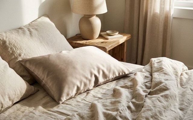 Pillow in a taupe silk pillowcase on a bed with beige linen sheets beside a ceramic lamp