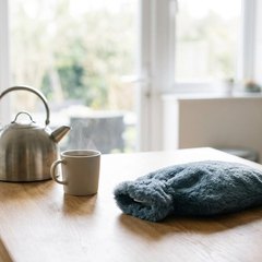 Hot water bottle in a soft blue-grey cover on a kitchen table beside a steel kettle and a mug of tea