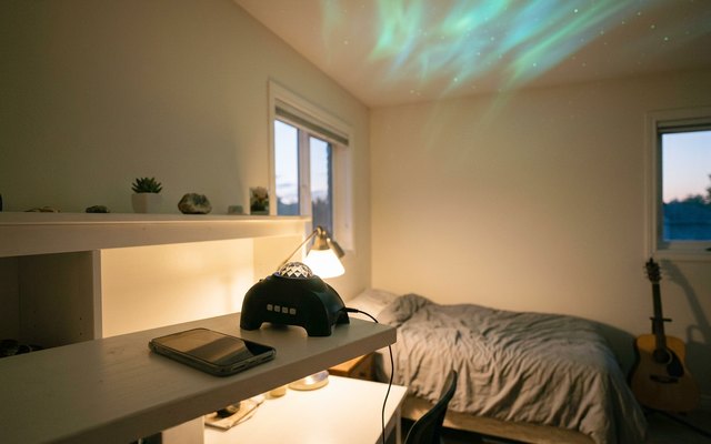 A small black aurora projector beside a phone on a desk shelf in a teenager’s bedroom, green and teal northern lights across the ceiling, a guitar by the bed