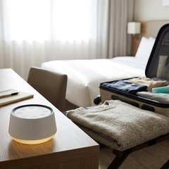 A small round white sound machine with a soft glow at its base on a hotel desk, beside an open suitcase and a folded sweater