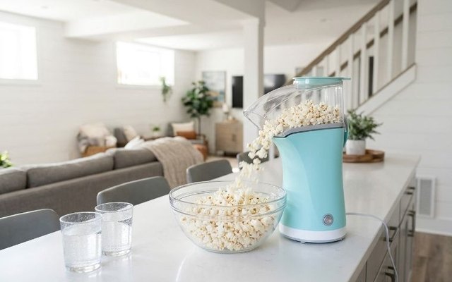 Aqua hot-air popcorn popper filling a glass bowl on a white bar counter in a bright basement rec room