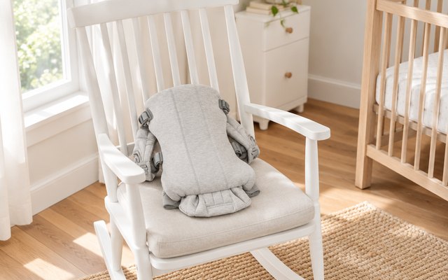 BabyBjorn Baby Carrier Mini, empty and laid flat on the seat of a rocking chair in a bright nursery