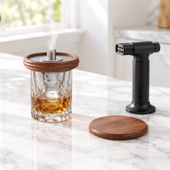 Mahogany cocktail smoker top on an old fashioned with a wisp of smoke, torch beside, on a white marble kitchen island