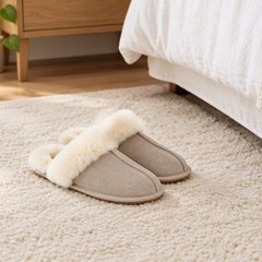 Sand suede slippers with a cream sheepskin collar, side by side on a cream rug beside a bed