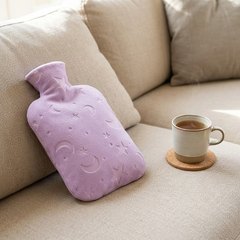 Hot water bottle in a lilac cover with moons and stars, resting on a sofa beside a mug of tea