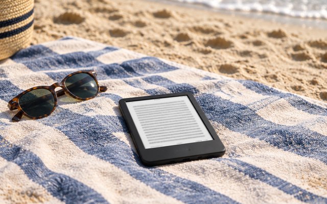 Kindle e-reader, on a striped beach towel on the sand beside a pair of sunglasses