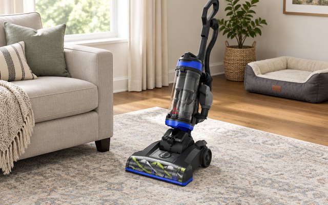 Bissell CleanView Swivel Pet vacuum, standing on a living room rug beside a sofa, an empty dog bed in the corner