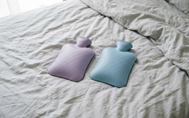 Two hot water bottles, one lilac and one blue, lying side by side on a rumpled grey bedsheet
