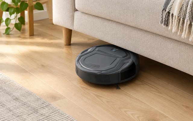 Small grey robot vacuum sliding under a low sofa on a light oak floor