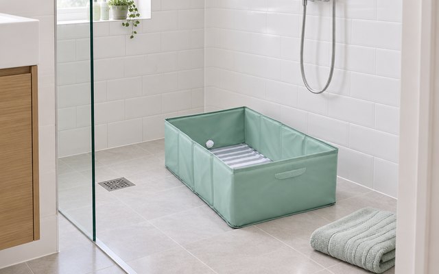 Ingenuity toddler tub, on the floor of a walk-in shower in a British bathroom with white tiles, a folded towel nearby