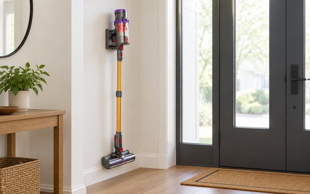 Gold and black cordless stick vacuum standing in its wall mount in a hallway beside a front door mat