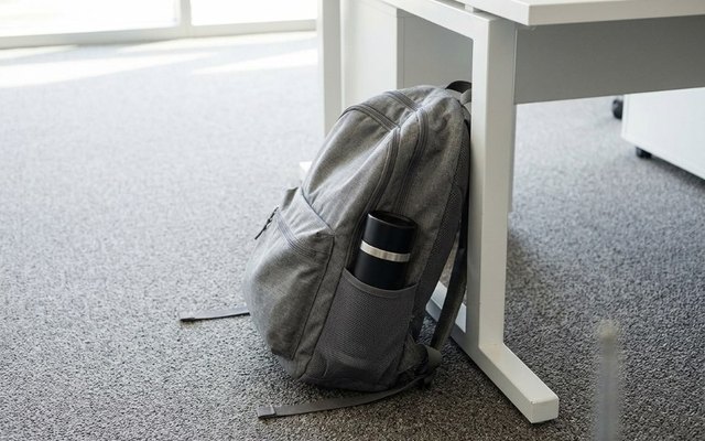 Small black travel bottle in the side pocket of a grey backpack by an office desk