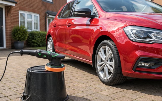 BATOCA T1 dual action polisher, standing on an upturned bucket beside a glossy red hatchback on a driveway in front of a brick semi-detached house