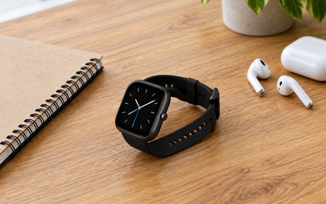 Black square smartwatch lying on a wooden desk beside a notebook and a pair of earbuds