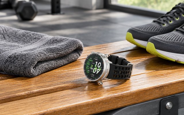 A round silver sports watch on a gym bench beside a towel