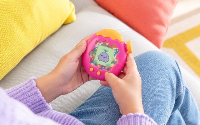 Pink egg-shaped digital pet with an orange dial held in a child's hands on a sofa