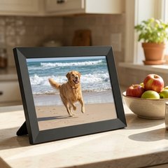 A black 10-inch digital photo frame on a sunny kitchen counter showing a dog running on a beach, beside a bowl of apples and a mug