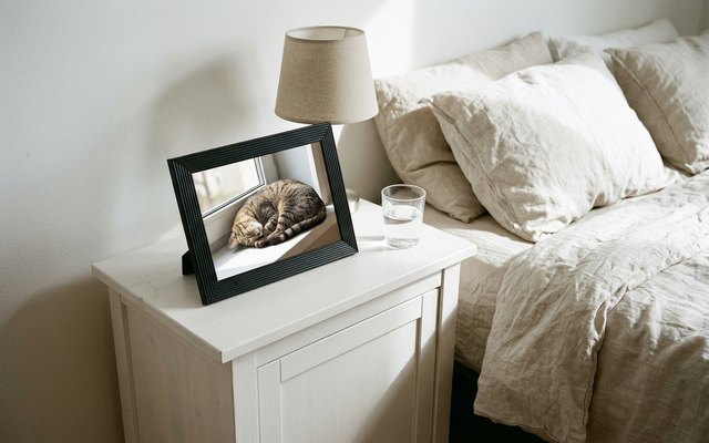 Black ridged digital photo frame on a white bedside table beside a lamp, showing a tabby cat asleep on a windowsill