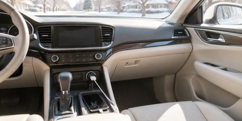 A small black car charger in the console socket of a sunlit car, a cable running to a phone in the tray, a snowy street outside