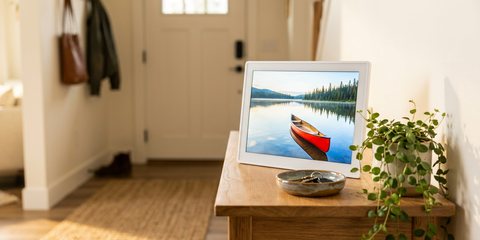 A white digital photo frame showing a red canoe on a lake, on an entryway console table beside a dish of keys and a trailing plant