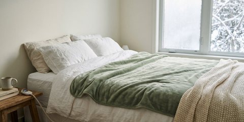 A sage-green heated blanket on a bed, its controller glowing on the nightstand, snowy trees outside the window