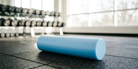 A smooth light blue foam roller on the rubber floor of a bright gym, a dumbbell rack and tall windows behind