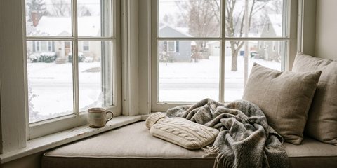 A hot water bottle in a cream cable-knit cover under a wool throw on a window seat, snow falling outside