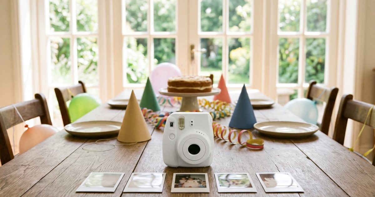 A white instant camera on a birthday party table with a row of fresh prints in front of it