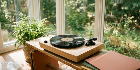 A light wood turntable playing a record on a low bench in a bright sunroom, beside a fern and a stack of record sleeves