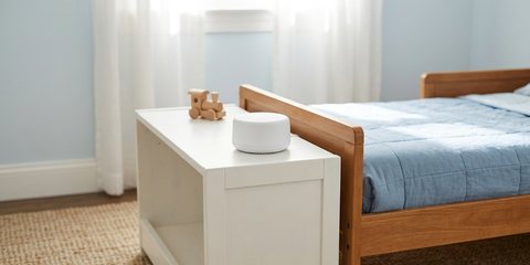 A small white sound machine on a low white shelf beside a toddler bed with a blue quilt, a wooden toy train next to it, in a bright bedroom