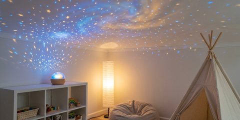 A small star projector on a low toy shelf in a kids’ playroom, casting blue and gold stars across the flat ceiling, a paper floor lamp and a teepee beside it