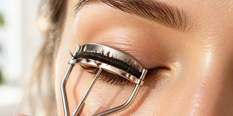 Extreme close-up of one closed eye with long natural lashes as a slim metal eyelash curler gently curls them, glowing skin, soft natural window light