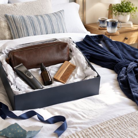 A just-opened birthday gift box of men's grooming things on a bed, with wrapping paper and a navy dressing gown