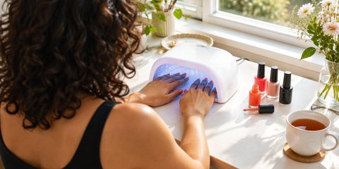 A woman seen from over her shoulder curing freshly painted gel nails in a white UV nail lamp